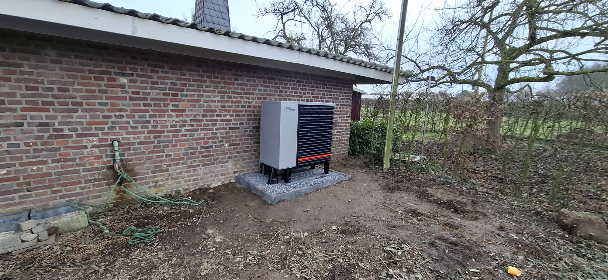 Außeneinheit einer Wärmepumpe der Marke alpha innotec auf Schotterfundament vor einer Backsteinwand installiert.